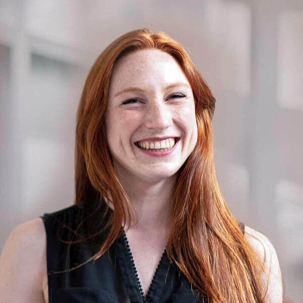 Eine lächelnde Frau mit langen, roten Haaren trägt ein schwarzes Oberteil und steht vor einem unscharfen Hintergrund.