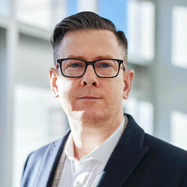 Ein Mann mit Brille und Anzug blickt selbstbewusst in die Kamera, in einem modernen, lichtdurchfluteten Büro.