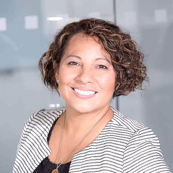 Eine lächelnde Frau mit lockigen Haaren trägt eine gestreifte Jacke und steht vor einem modernen Bürohintergrund.