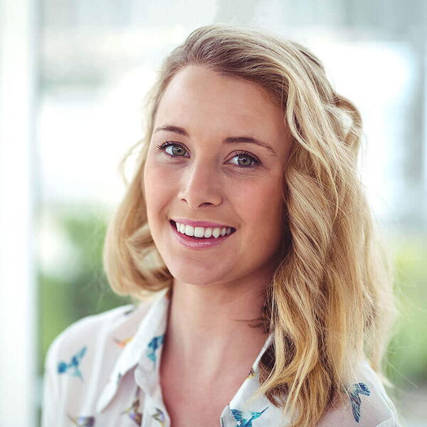 Eine lächelnde Frau mit blonden Haaren trägt eine Hemdbluse mit Vogelmuster vor unscharfem Hintergrund.